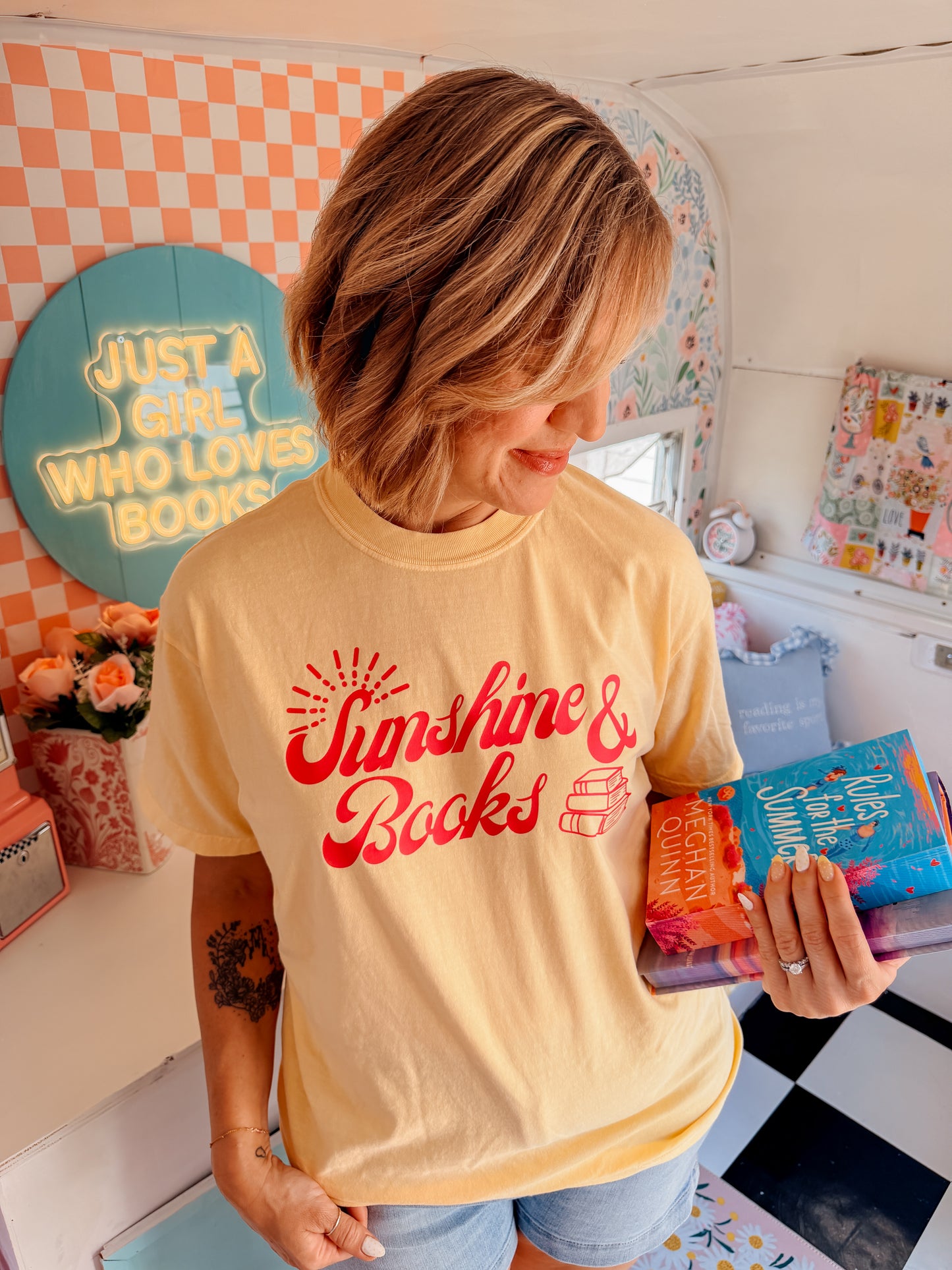 A woman wearing a pale yellow graphic t-shirt with red text reading 'Sunshine & Books' and holding a colorful book inside a decorated room with book-themed decor.