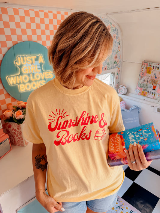 A woman wearing a pale yellow graphic t-shirt with red text reading 'Sunshine & Books' and holding a colorful book inside a decorated room with book-themed decor.