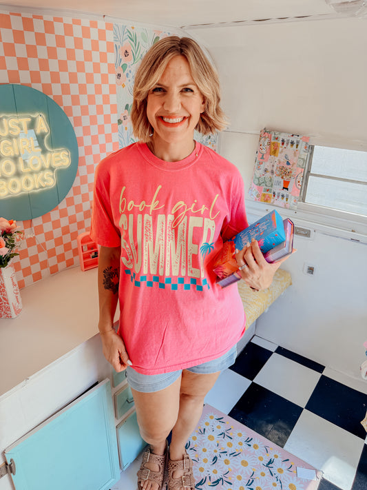 A woman stands in a retro-themed room wearing a pink graphic t-shirt that reads 'book girl SUMMER' with a blue and pink checkerboard stripe design. She is holding a colorful book or notebook.