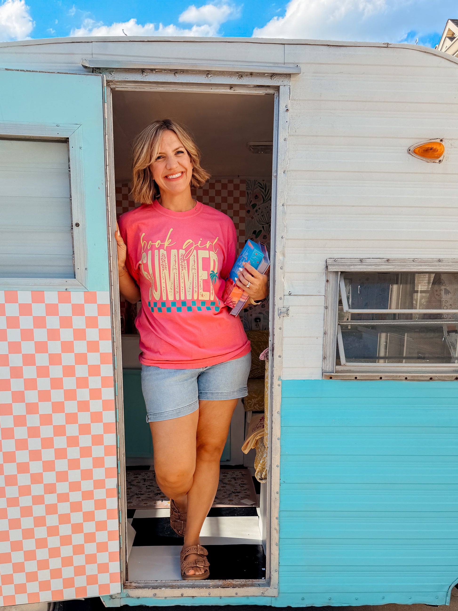 A woman stands in the doorway of a pastel blue and white trailer wearing a pink graphic t-shirt that reads 'Luv girl SUMMER' and denim shorts, holding a blue bottle.