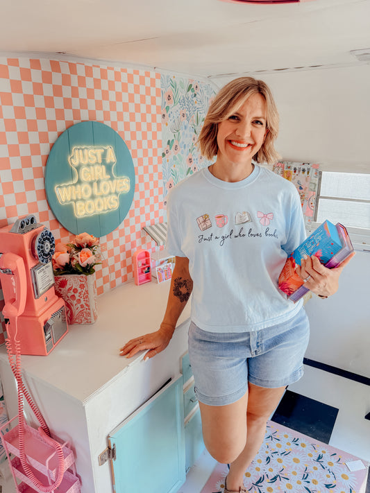 A woman stands in a retro-themed room wearing a light blue graphic t-shirt and denim shorts, holding a pink and blue book. The background features pink and white checkered wallpaper, a vintage pink rotary phone, and a sign that reads 'JUST A GIRL WHO LOVES BOOKS'.