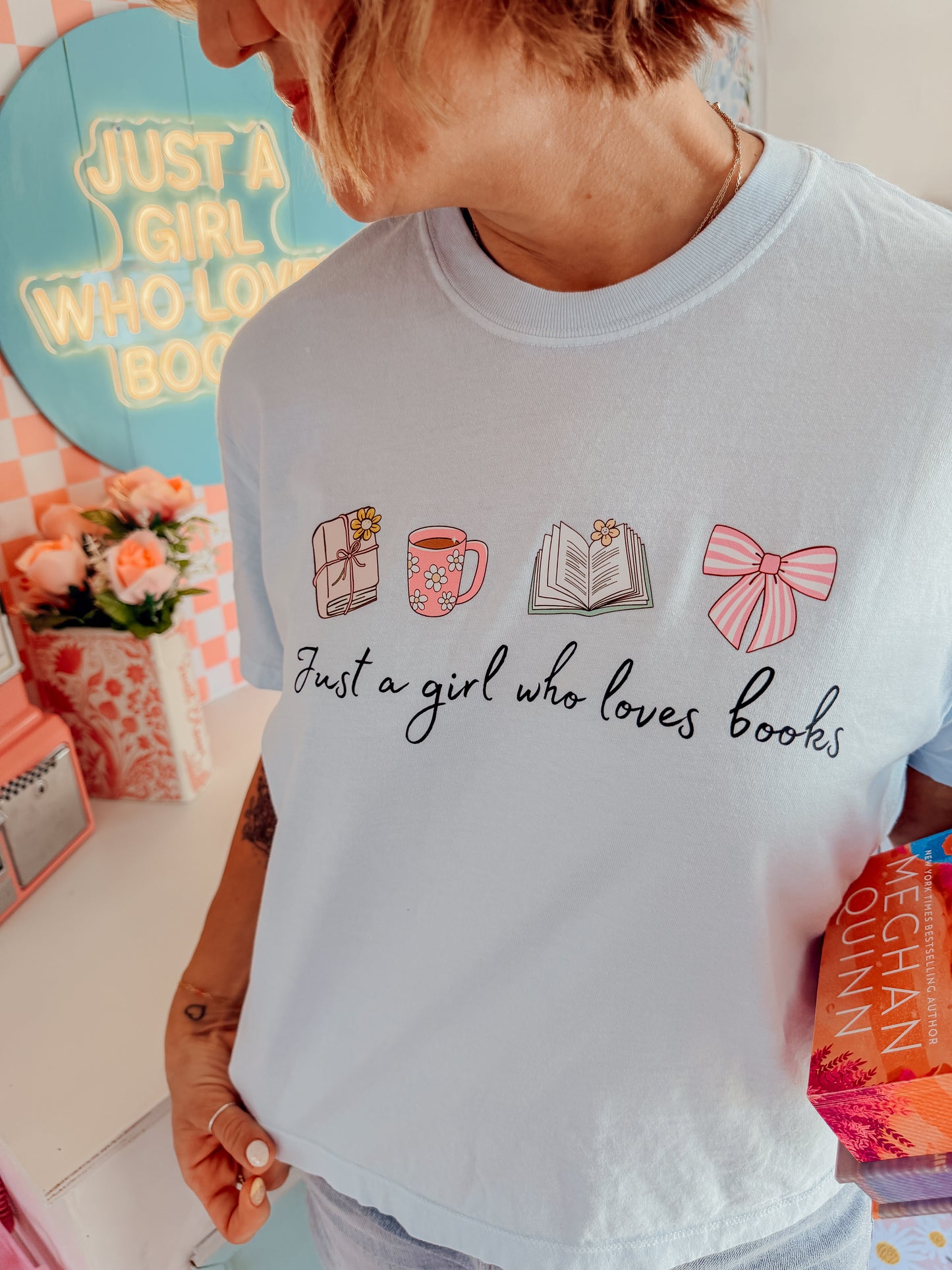A woman wearing a light blue graphic t-shirt with illustrations of a gift box, a pink mug, an open book, and a pink bow, with the text 'Just a girl who loves books'.