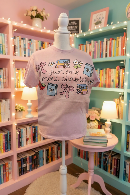 A light pink graphic t-shirt displayed on a white dress form mannequin in a cozy, colorful bookshop setting. The t-shirt features a whimsical illustration of books, flowers, and bows with the text 'Just one more chapter!'.